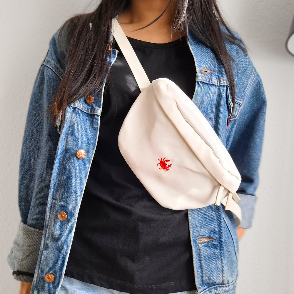 Model trägt weisse Bauchtasch über den Körper. Auf der Tasche ist eine kleine rote Krabbe darauf gestickt.