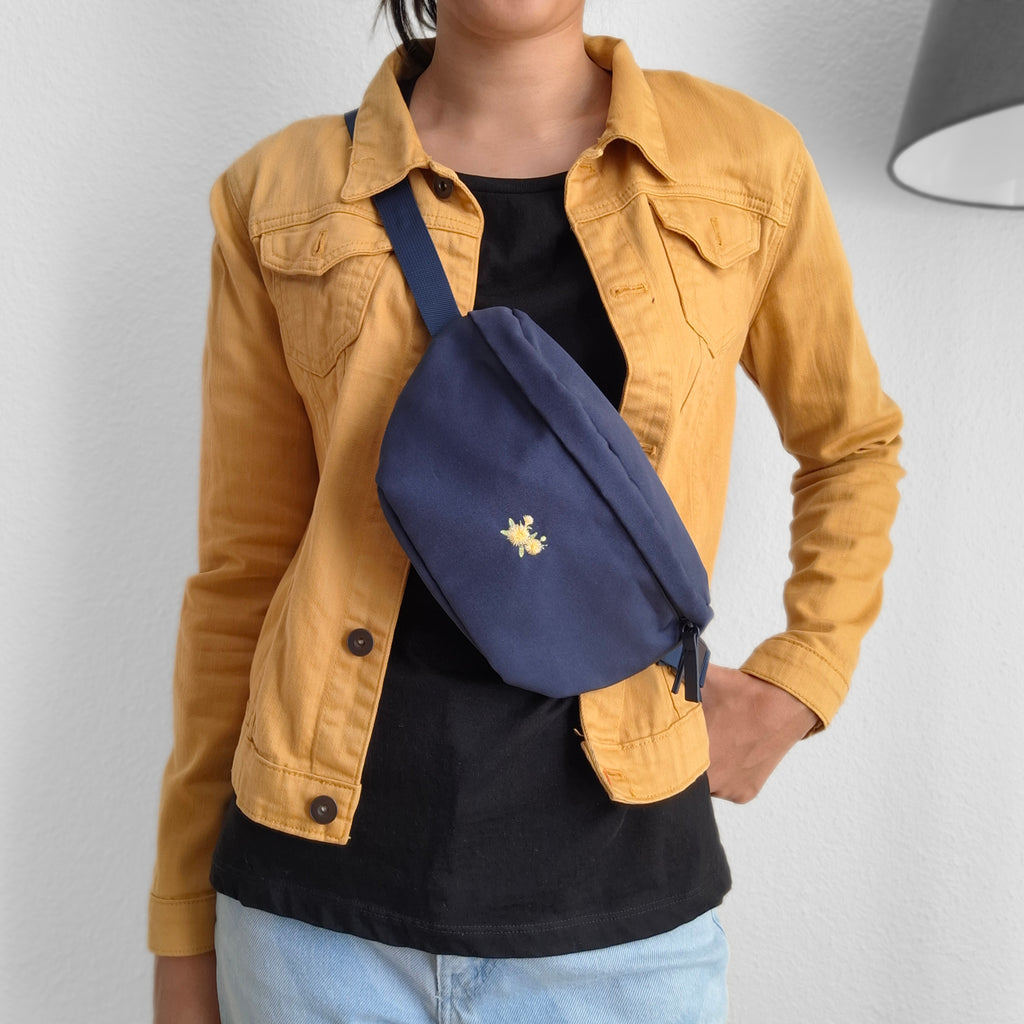 Model trägt dunkelblaue Bauchtasche über die Schulter. Auf der Tasche ist eine Löwenzahn-Pflanze darauf gestickt.