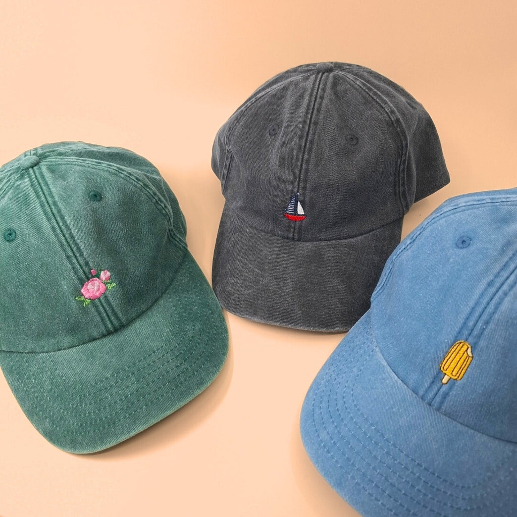 Drei Caps mit bestickten Icons. Von links nach rechts: Grüne Kappe mit Rose, schwarze Kappe mit Segelboot, blaue Cap mit Eis am Stiel.