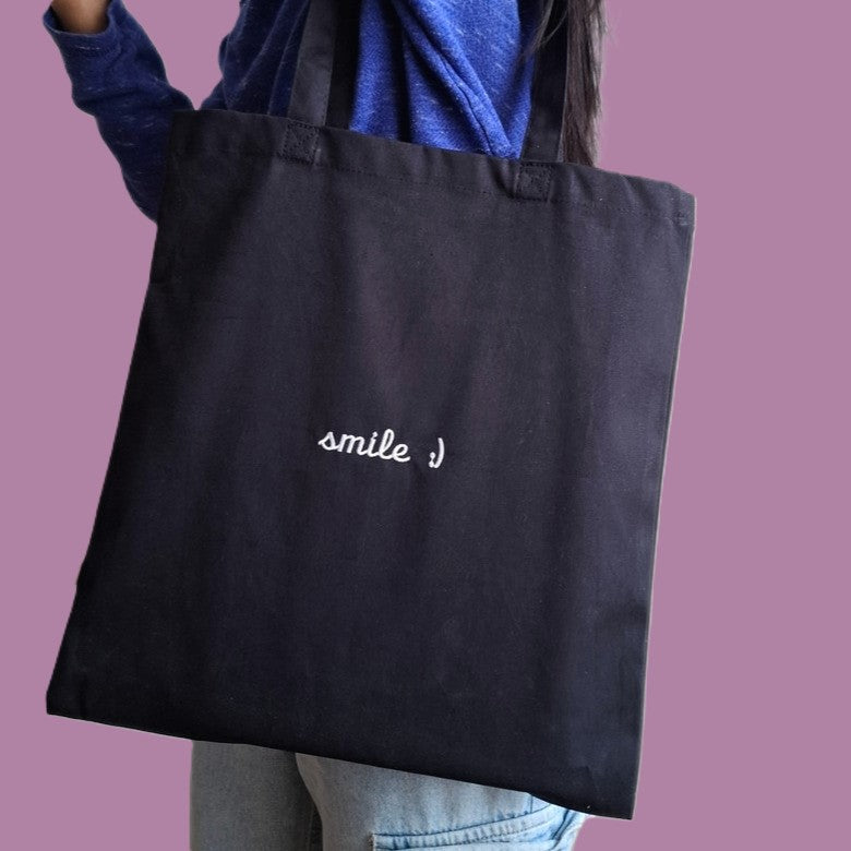 Person trägt eine schwarze Stofftasche mit dem Text "smile :)" darauf gestickt in weiss.