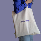 Person trägt eine beige Stofftasche mit dem Text "alles easy" darsuf gestickt in violett