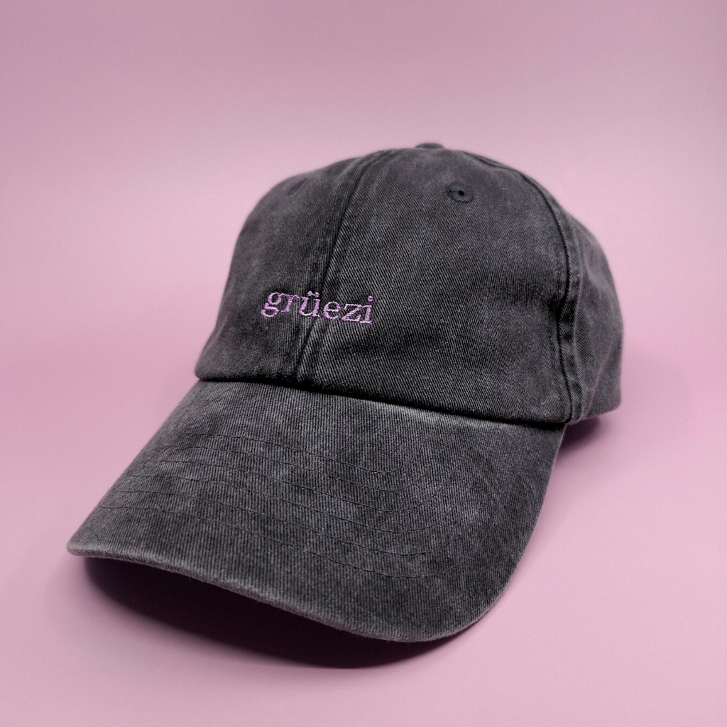 Schwarze Cap mit besticktem Text "grüezi" in lila und einer Serifen-Schrift.