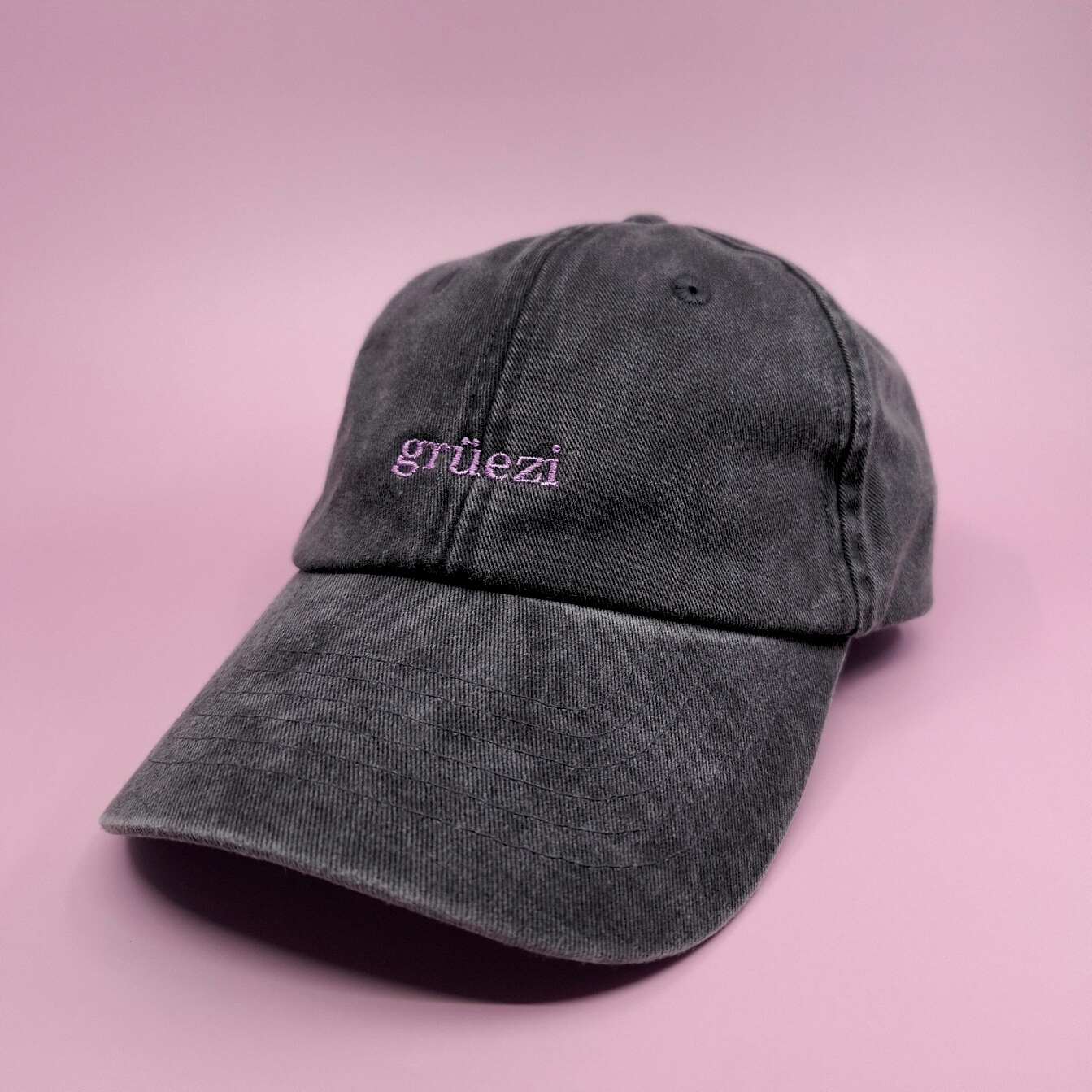Schwarze Cap mit besticktem Text "grüezi" in lila und einer Serifen-Schrift.