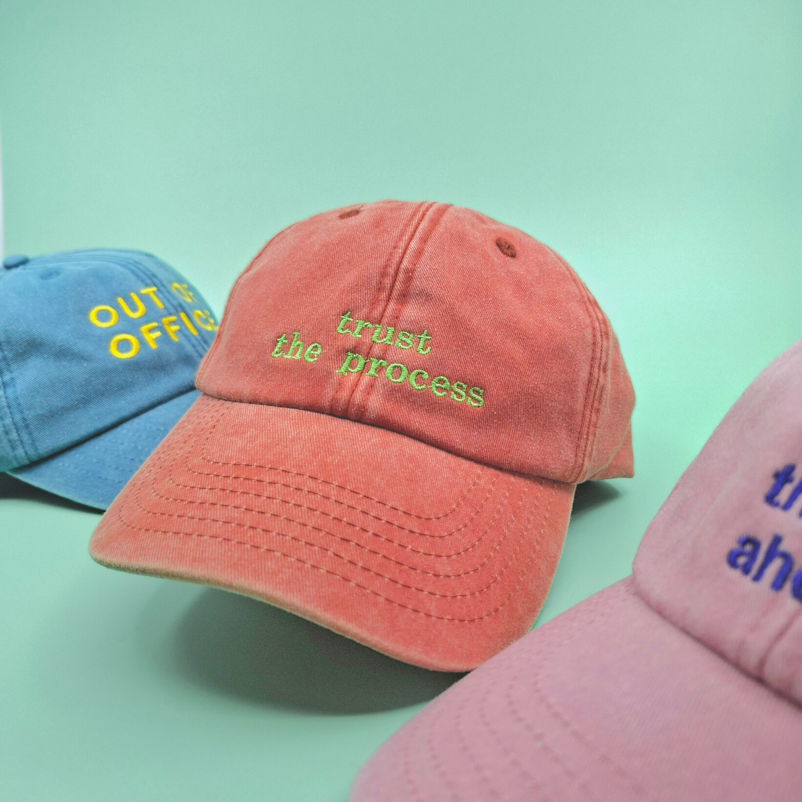 Drei farbige Caps. Im Mittelpunkt eine orange Cap mit dem Schriftzug "trust the process" in grün und einer Serifen-Schrift.