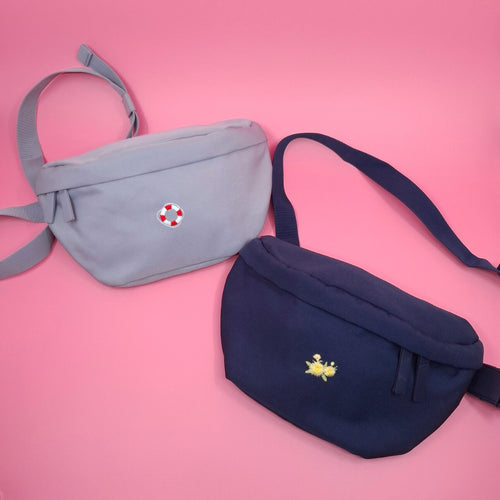 Eine blau-graue Bauchtasche mit einem Rettungsring-Icon darauf gestickt und eine dunkelblaue Tasche mit einem Löwenzahn-Icon.